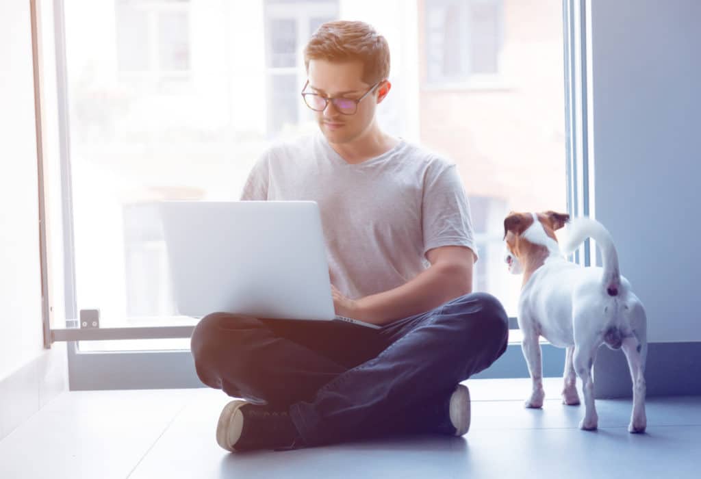 Man sitting cross-legged on the floor with his laptop while his dog looks out the window.