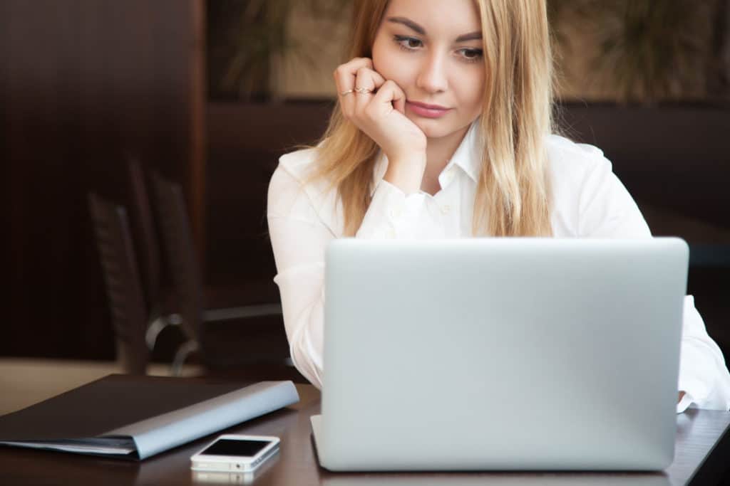 Young beautiful blond caucasian business woman working on laptop with concentrated, serious expression, cell phone and black dossier lying on the table