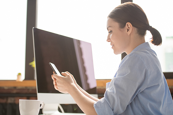 Young casual female employee looking at smartphone screen while scrolling and searching for message or information