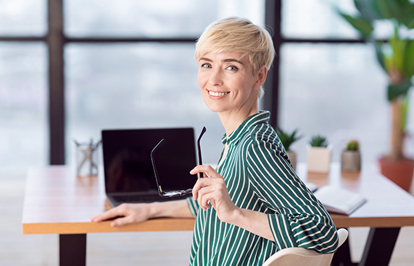 Smiling Mature Businesswoman Looking At Camera Posing Holding Eyeglasses Sitting In Modern Office. Selective Focus