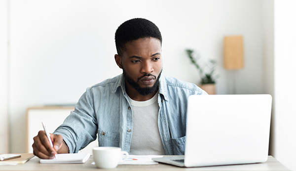 Job search. Unemployed man using laptop searching for vacancies online in internet and taking notes, panorama with copy space/ wearing blue jean shirt